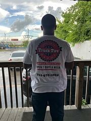 A person stands with their back to the camera, wearing a white T-shirt with "Truck Tire" branding and contact information, promoting tire services.