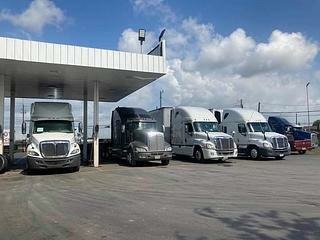A row of semi trucks parked at a fuel station under a blue sky, showcasing various truck models and colors, highlighting transport logistics.